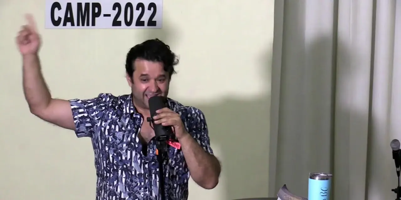 Un predicador habla con el micrófono y levanta la mano bajo un letrero que dice «CAMP-2022», en la bienvenida de REBORN 2022.