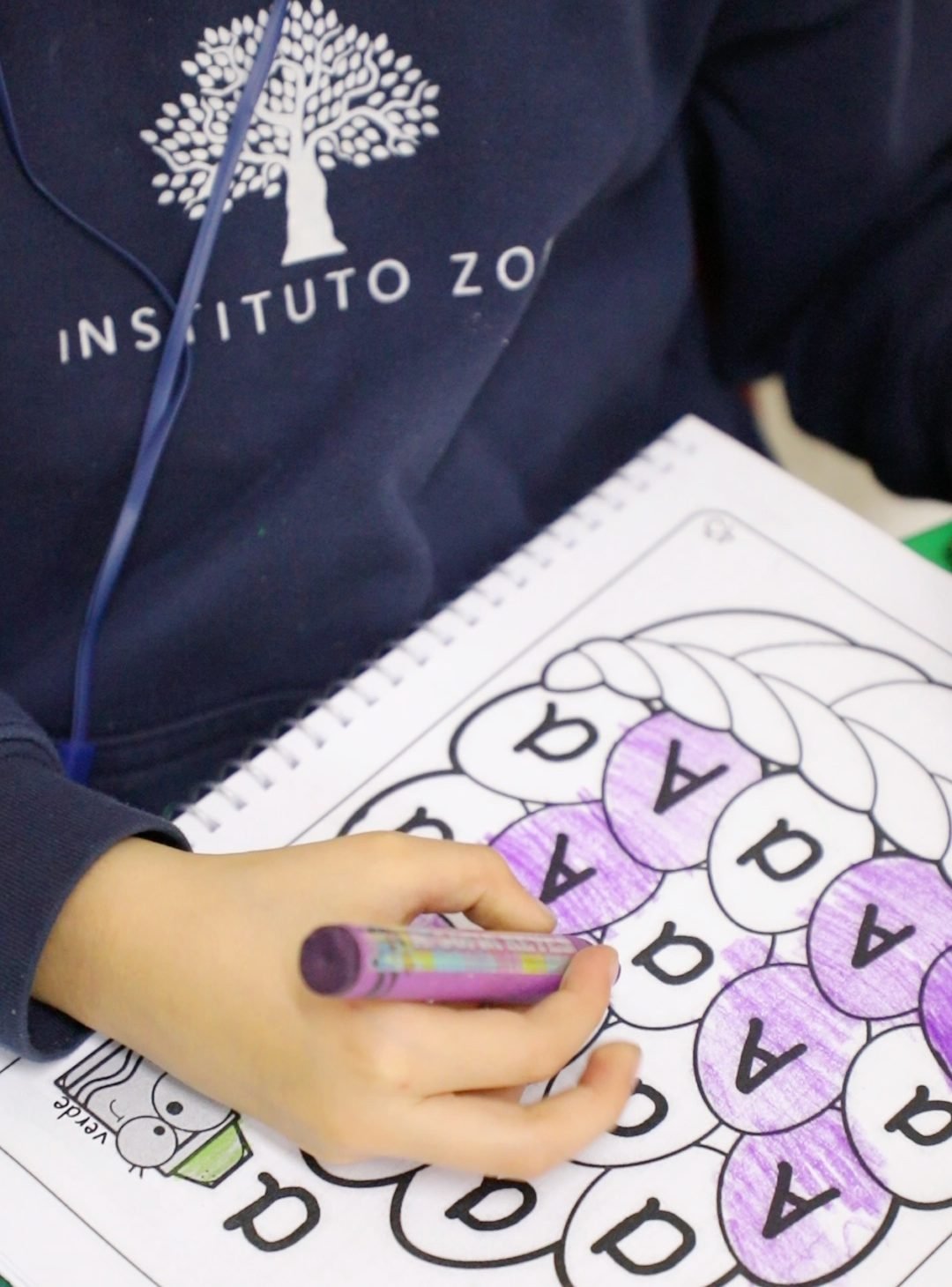 La mano de una alumna del Instituto Zoe colorea de morado las letras «A» de su hoja de trabajo