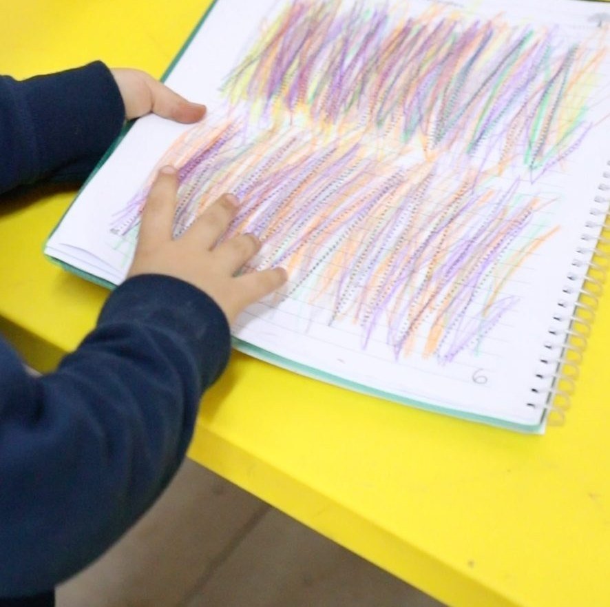 Una mano llena de trazos de colores su hoja de trabajo, sobre la mesa amarilla del salón