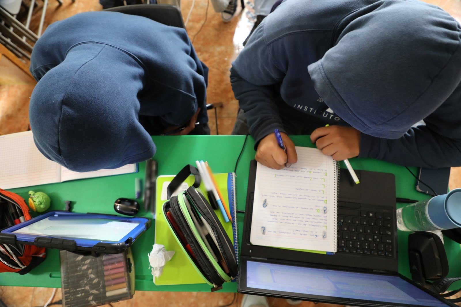 Dos alumnos de secundaria del Instituto Zoe, con la capucha de la sudadera puesta, escriben en su cuaderno sobre la mesa verde, entre su laptop, una tableta y sus útiles; vista desde arriba